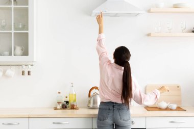 Genç milenyum ev kadınlarının arka arkaya bakış açısı, turuncu egzoz kaputunu seçen ve seçen mod, ocağın üstündeki mekanik vantilatör düğmesine basan, modern mutfakta evde duran