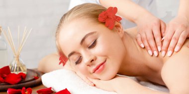 Rahatla, iyileşme ve güzellik. Gülümseyen kız çiçek yaprakları, aromatik çubuklar ve yağ ile masaj masasında yatıyor.