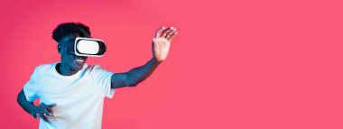 Genç siyahi bir adam tamamen sanal gerçeklik deneyimine dalmış, kulaklık takıyor ve görünmeyen dijital bir çevreyle etkileşime girerken geniş bir gülümseme sergiliyor, canlı pembe bir arka planla çevrili..