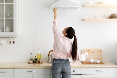 Ev kavramı. Arka planda, sıradan bir bayanın menzil egzoz kaputunda seçtiği ve seçtiği mod, ocağın üstündeki mekanik vantilatörün düğmesine basıyor. Evdeki modern mutfakta yemek pişiren kadın.