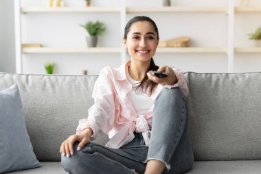 Boş zaman ve eğlence. Televizyon programı ya da film izleyen, uzaktan kumandayla gülümseyen genç bir kadın. Oturma odasındaki rahat koltukta oturup eğlenen neşeli bayan.
