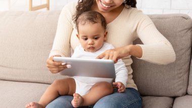 Gülümseyen Afrikalı anne, bebeğini kucağında tutuyor, dijital tablet kullanıyor. Bebeklerin eğitimini bilgisayarda gösteriyor, öğreniyor ve evdeki koltukta otururken eğleniyor. Aile Ağı, Teknoloji