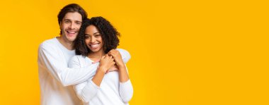 Mutlu ırklar arası çift portresi. Neşeli melez sevgililer kameraya sarılıyor ve poz veriyor, sarı arka planda yan yana duruyorlar, panorama
