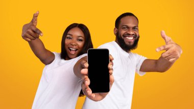 Yeni Mobil Uygulama Reklamı. Elinde boş ekranla akıllı telefonu tutan mutlu siyah çiftin portresi, model için boş alanı olan cihazları gösteriyor ve işaret ediyor, sarı stüdyo arka planı