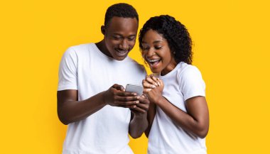 Heyecanlı siyah çift cep telefonu ekranına bakıyor, yeni bir uygulama kullanıyor, sarı stüdyo geçmişi var. Neşeli Afrikalı Amerikalı erkek ve kadın akıllı telefon kullanıyor, eğlence konsepti.