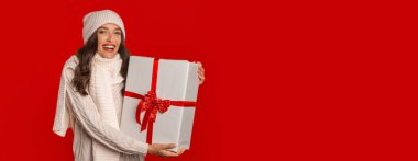 Noel Baba şapkası takan neşeli milenyum bayanı kırmızı stüdyo arka planında Noel beklentisiyle hediye kutusunu tutuyor. Tatil sezonu reklamları için mükemmel bir pankart, kopyalama alanı