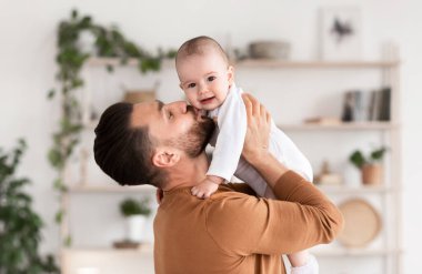 Babalar sever. Sevimli, genç bir babanın küçük kızını öpmesi ve kucağına alıp kucağına alması kucağında kucağında yeni yürümeye başlayan bebekle evde durması. Ebeveynlik ve Babalık Keyfi