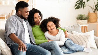 Küçük kızı olan modern Afrikalı Amerikan ailesi evde birlikte vakit geçiriyorlar, koltukta dinleniyorlar ve sarılıyorlar.