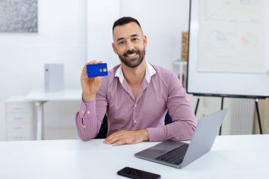Elinde mavi kart olan sakallı bir adam gülümsüyor. Bilgisayarı ve akıllı telefonu olan bir masada oturuyor. Bu da profesyonel bir ortam ve ilgi çekici bir an olduğunu gösteriyor..