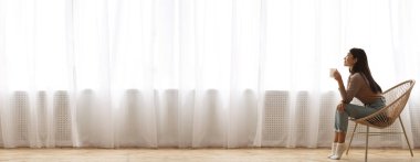 Günaydın. Rahatla. Geniş açıda, hasır sandalyede oturan ve pencerenin önünde kahve içen Asyalı bir kız var.