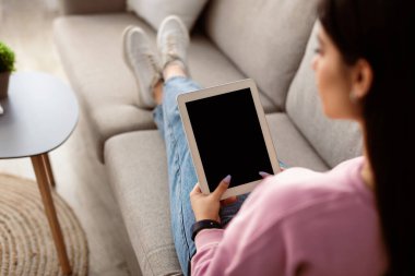 Omuz hizasında, dijital tabletli genç bir kadının şablon oluşturmak için siyah boş ekranı tutması, koltukta oturması, elektronik kitap okuması ya da video araması yapması.