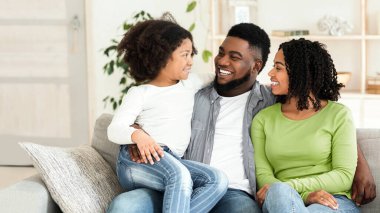 Aile zamanı. Mutlu Afrika kökenli Amerikalı ebeveynler evlerinde küçük kızlarıyla kaynaşıyor, birlikte oturma odasında oturuyor ve gülümsüyorlar.