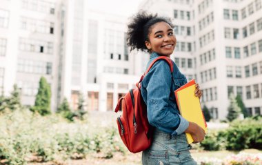 Sırt çantalı, gülümseyen kitaplarla poz veren Afro-Amerikalı Öğrenci Kız Açık Üniversite Binası 'nın yanında duruyor. Modern Eğitim ve Öğrencilik Yaşam Tarzı