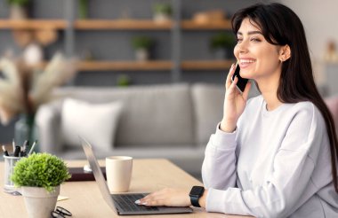 Telekomünikasyon konsepti. Cep telefonuyla konuşan neşeli bir kadının portresi, masada oturup evde dizüstü bilgisayarda çalışan neşeli serbest çalışan bir kadının bilgisayar kullanması ve hoş bir cep telefonu konuşması yapması.