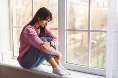 Akıl Sağlığı Konsepti. Üzgün genç bir kadın pencerenin kenarında oturuyor, bacaklarını kucaklıyor ve pencereden dışarı bakıyor. Yalnız kadın depresyonda, yalnız hissediyor, umutsuz ya da melankolik.