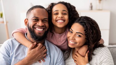 Sevgi ve ilgi. Evdeki kanepede oturup fotoğraf için poz veren ve kameraya bakan üç kişilik neşeli Afro-Amerikan aile portresi. Gülümseyen genç kız ailesini kucaklıyor.