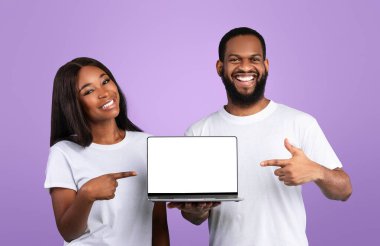 Şu siteyi kontrol et. Siyah ekranlı dizüstü bilgisayarı tutan mutlu Afrikalı erkek ve kadın portresi, modelleme için boş alanı olan bilgisayarları işaret ediyor, mavi stüdyo arkaplan, afiş.