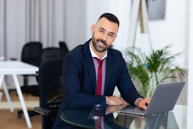 Profesyonel bir adam, modern bir ofiste masasında oturuyor. Dizüstü bilgisayarıyla meşgul. Kendine güvenen, takım elbiseli, minimalist ve iyi tasarlanmış bir ortamda gülümsüyor..