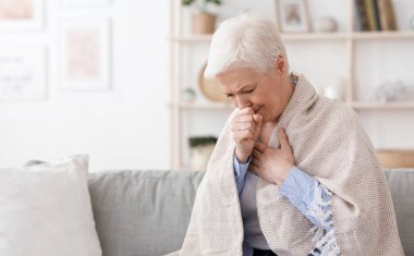 Yaşlı kadın battaniyeye sarılmış, evde öksürüyor, Coronavirüs belirtileri gösteriyor, oturma odasında koltukta oturuyor, boşluğu kopyalıyor.