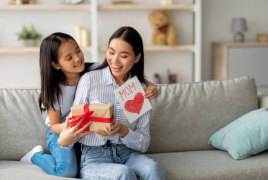 Kutlarım. Sevgi dolu Asyalı kız, Anneler Günü 'nde anneye sürpriz yaptı. El yapımı kart ve hediye kutusu verdi. Ev içi koltukta oturdu.
