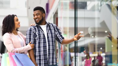 Sevgilisiyle alışveriş yapan, vitrindeki yeni kıyafetlere işaret eden, indirimli fiyatlar gösteren, sezonluk satışların keyfini çıkaran genç Afrikalı Amerikalı çift,