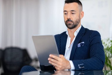 İnce çizgili takım elbiseli iyi giyimli bir adam çağdaş bir ofiste gösterişli bir masada otururken tabletine odaklanır. Doğal ışık büyük pencerelerden süzülerek profesyonel atmosferi geliştirir..