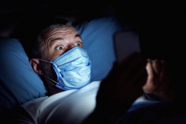 Coronavirus. Çevrimiçi haber okuyan, geceleri yatakta yatan, tıbbi maske takan, akıllı telefon kullanan korkmuş bir adam. Panorama
