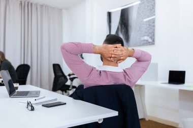 Sıradan gömlekli bir adam, elleri başının arkasına sıkıca bağlanmış, masa ve dizüstü bilgisayarlarla çevrili, çağdaş bir ofis ortamında bir anlık rahatlığın tadını çıkarıyor..