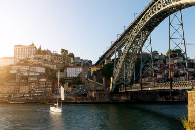 Bir yelkenli Porto 'da çarpıcı bir köprünün altındaki sakin sularda zarifçe hareket eder. Gün batımı, canlı evlerin üzerine sıcak bir parıltı salıyor. Rıhtım duvarını kaplıyor. Resmedilmeye değer bir sahne oluşturuyor..