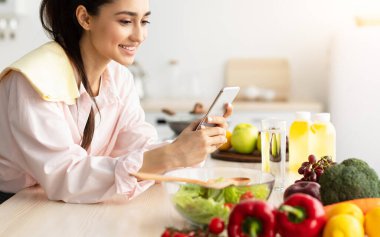 Mutfakta yemek pişirirken cep telefonu kullanan gülümseyen genç bir kadın masaya yaslanan neşeli bir kadın cep telefonundan mesajlar okuyan ve gülümseyen, bedava fotokopi çeken bir kadın.