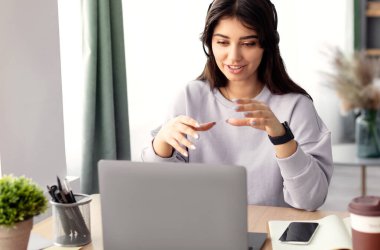Çevrimiçi Video Çağrısı. Masada oturmuş gülümseyen bir kadının portresi dizüstü bilgisayarda sanal konferans yapıyor, web kamerasına konuşuyor ve el kol hareketi yapıyor. İletişim Konsepti. Bilgisayar kullanarak kulaklık sohbeti yapan bir kadın.