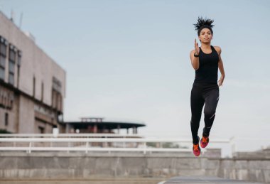 Running, jump and flight. Athletic slim african american girl in fashionable sport uniform froze in air on street against sky background, free space clipart