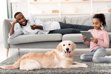 Mutlu siyahi aile portresi birlikte zaman geçiriyor, kızı köpekle yerde oturuyor ve tablet kullanıyor, babası oturma odasında kanepede uzanıyor, uzaktan kumandayı elinde tutuyor.