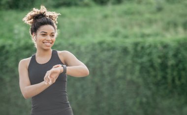 Healthy lifestyle and heart rate check. Smiling slim african american girl in sportswear with wireless headphones looking at fitness tracker in park, sun flare, free space clipart