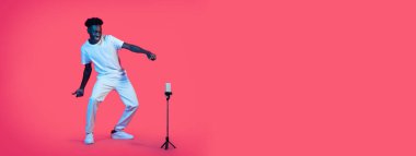 Beyaz tişört ve beyaz pantolon giyen siyah bir adam bir standın önünde telefonun önünde dans ediyor. Kollarını uzattı ve gülümsüyor. Arka plan parlak pembe..