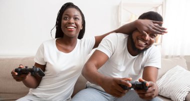 Fans Of Game Console. African American Couple Playing Videogames Sitting On Couch At Home, Woman Trying To Cover Boyfriends Eyes panorama