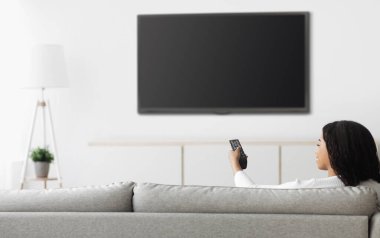 Afro-Amerikalı bir kadın televizyon izlerken düz ekran plazma televizyona uzaktan kumandayla işaret ederek boş ekran, evde koltukta otururken kanal değiştirerek, maket yaparak...