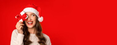 Küçük bir Noel hediyesi ile poz veren Avrupalı genç bir kadın, kırmızı stüdyo arka planında hediye kutusuyla gözlerini kapatıyor. Eğlence ve hediye verme zamanı geldi çattı. Panorama, boş alan