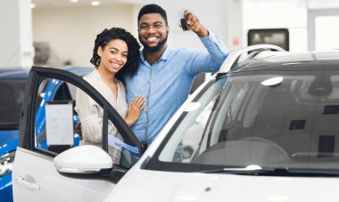 Cheerful Afro Spouses Standing Near New Car Showing Key Smiling To Camera In Auto Dealership Center.