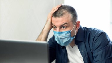 Coronavirus Haberleri. Evde oturan koruyucu tıbbi maske takan şok olmuş adam dizüstü bilgisayar kullanıyor.