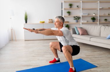 Çalışmak için evde tek başına kal. Spor kıyafetleri içinde olgun bir adam oturma odasının içinde çömelme yapıyor, koronavirüs izolasyonu sırasında ev içi eğitimlerde bacaklarını çalıştırıyor, serbest alan.