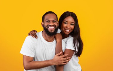 Aşık çift. Kameraya sarılıp poz veren neşeli siyah sevgililerin portresi. Stüdyoda sarı arka planda izole bir şekilde dikiliyorlar. Kopya alanı. Sıradan bir adam ve kadın gülümsüyor.