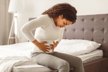 Portrair of upset black woman suffering from strong abdominal pain, touching her tummy, sitting on bed. Sad young lady feeling acute stomachache, free copy space. Chronic disease, spasm, hernia clipart