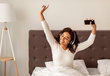 Kulaklığı ve cep telefonu olan mutlu siyah kadın evde sevdiği müzikle dans ediyor, yatakta oturuyor. Afro-Amerikalı kadın güzel bir şarkıya taşınıyor, sabahları harika vakit geçiriyor.