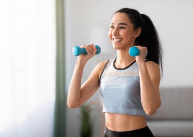 Evde dambıl egzersizi yapan, kol gücü üzerinde çalışan, fotokopi odasına bakan neşeli genç bir kadının yakın çekimi. Gülümseyen atletik kadın oturma odasının iç tarafına mavi fitness damlalarını kaldırıyor.