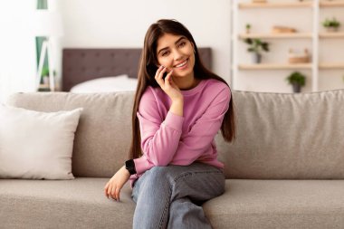 Pozitif Duygular Konsepti. Sıradan güzel bir genç kadının portresi gülümseyen, poz veren ve kameraya bakan, yanağa dokunan. Evdeki oturma odasında rahat koltukta oturan neşeli kadın manken.