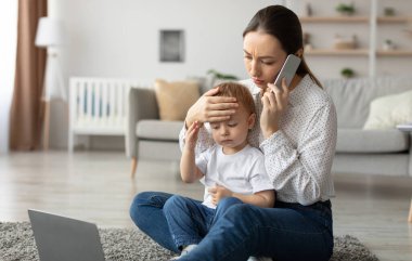 Call to pediatrician. Stressed mother talking to doctor, worried about her sons health, checking childs temperature by holding hand on kid forehead. Remote healthcare consultation.