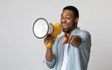 Pozitif Afro-Amerikalı adam megafonla bağırıyor ve gri arka plan üzerinden kameraya işaret ediyor, kopyalama alanı. Hoparlörle bağıran, neşelenen, reklam yapan mutlu siyah adam.