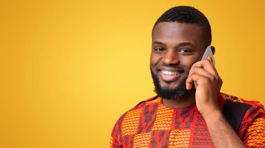 Geleneksel Afrika kıyafetleri içinde gülümseyen siyah adamın yakın plan sarı arka plan üzerinden cep telefonuyla konuşması, boşluğu kopyala