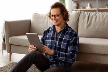 Mutlu Milenyum Adamı Evdeki Oturma Odasında Yerde Oturan Dijital Tableti Kullanıyor. Hippi Adam içeride bilgisayardan film izliyor. Gadget ve Teknoloji Konsepti